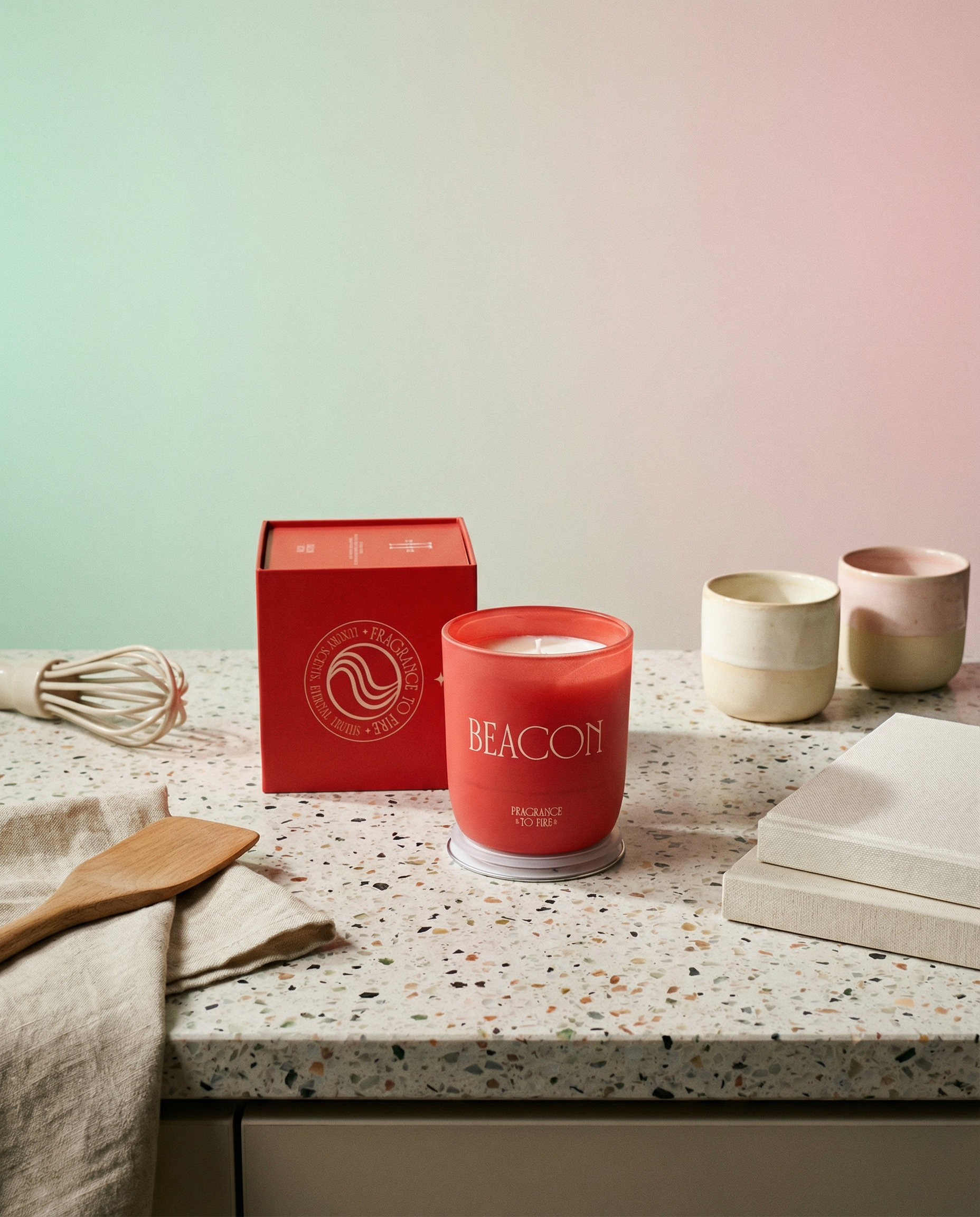 Red candle labeled 'Beacon' on a speckled countertop with a red box and kitchen utensils. Terracotta Beacon candle lit, calming aromatherapy candle for focus, clarity, and peace.