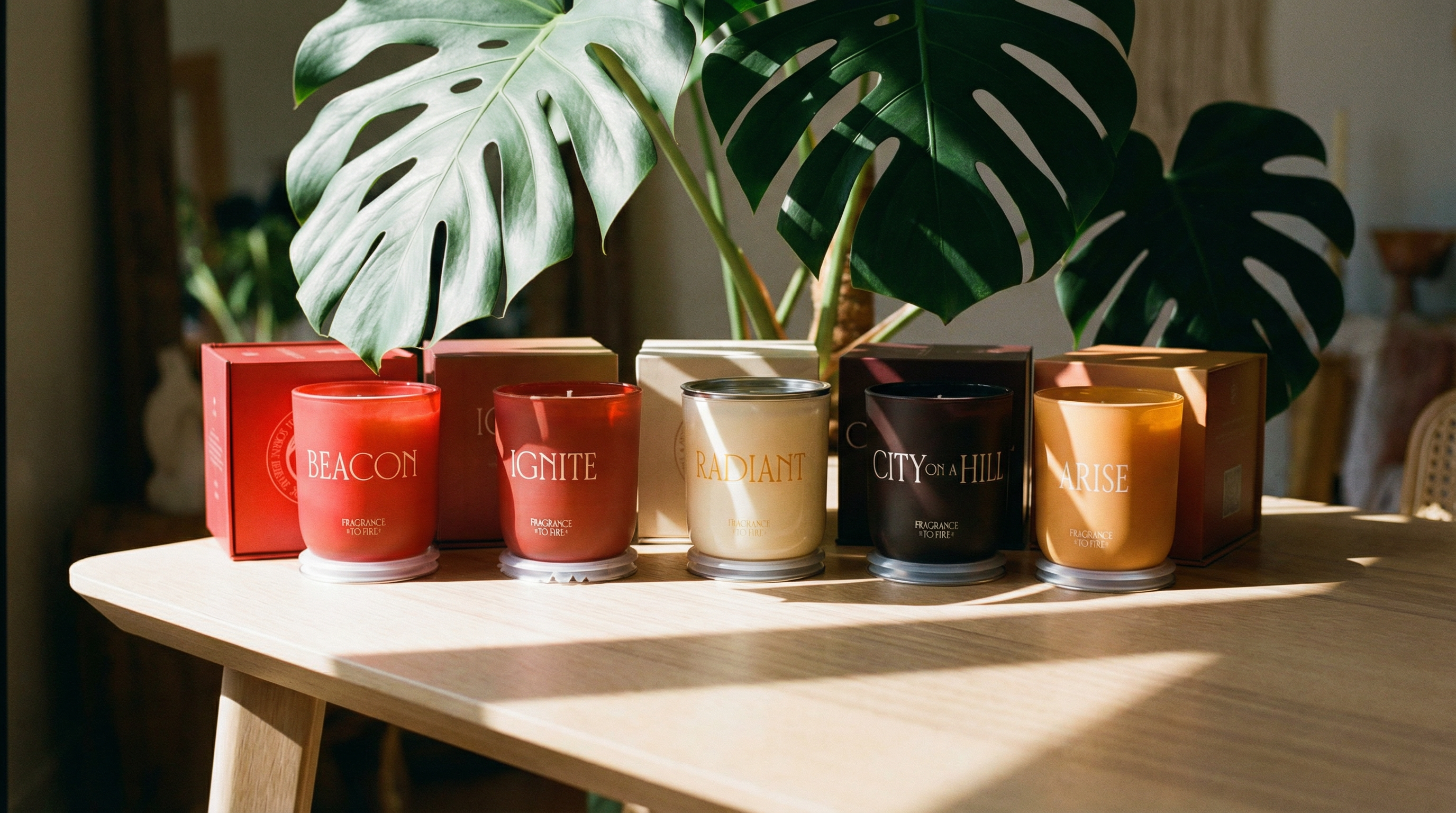 Row of candles with visible brand names on a wooden table with plants in the background